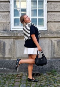 Bolso negro de forma redondeada con logo, zapatos planos de lunares, falda blanca y una camiseta negra con una bufanda a rayas caída sobre los hombros.