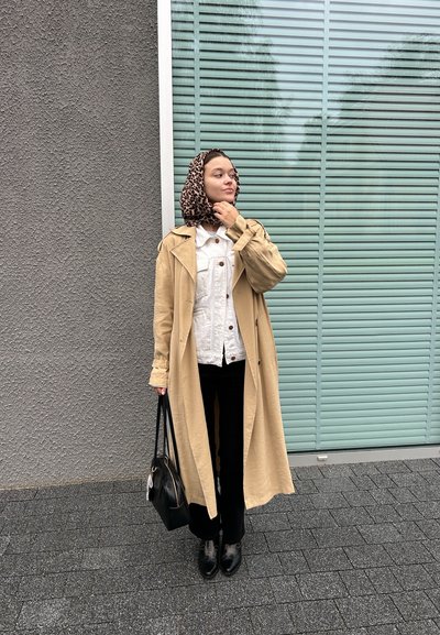 Gabardina beige sobre una camisa blanca abotonada, pantalones negros y zapatos negros. Pañuelo con estampado. Bolso negro. Fondo con persianas verdes.