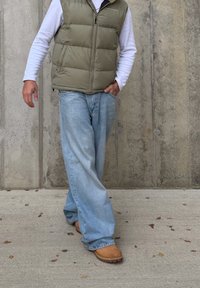 Persona che indossa jeans azzurri chiari, stivali color cuoio, camicia bianca a maniche lunghe e gilet imbottito verde oliva mentre cammina su una superficie di cemento accanto a un muro di cemento.