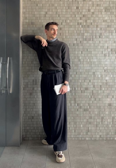 Modelo con un suéter de carbón y pantalones de pierna ancha negros, sosteniendo una carpeta blanca. De pie contra una pared de baldosas grises texturizadas.