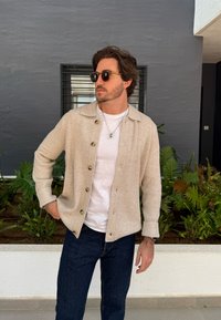 Bežs džemperis ar pogām priekšpusē, ar apkakles dizainu, apvienots ar baltu T-kreklu un tumši zilām džinsām. Aksesuāri ietver kaklarotu.