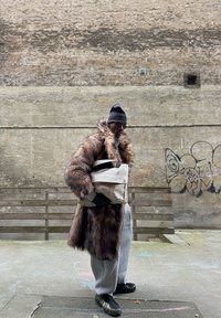 Manteau en fausse fourrure marron à manches longues, pantalon de survêtement gris, et sac beige. Accessoires : bonnet noir et lunettes de soleil. Arrière-plan urbain.