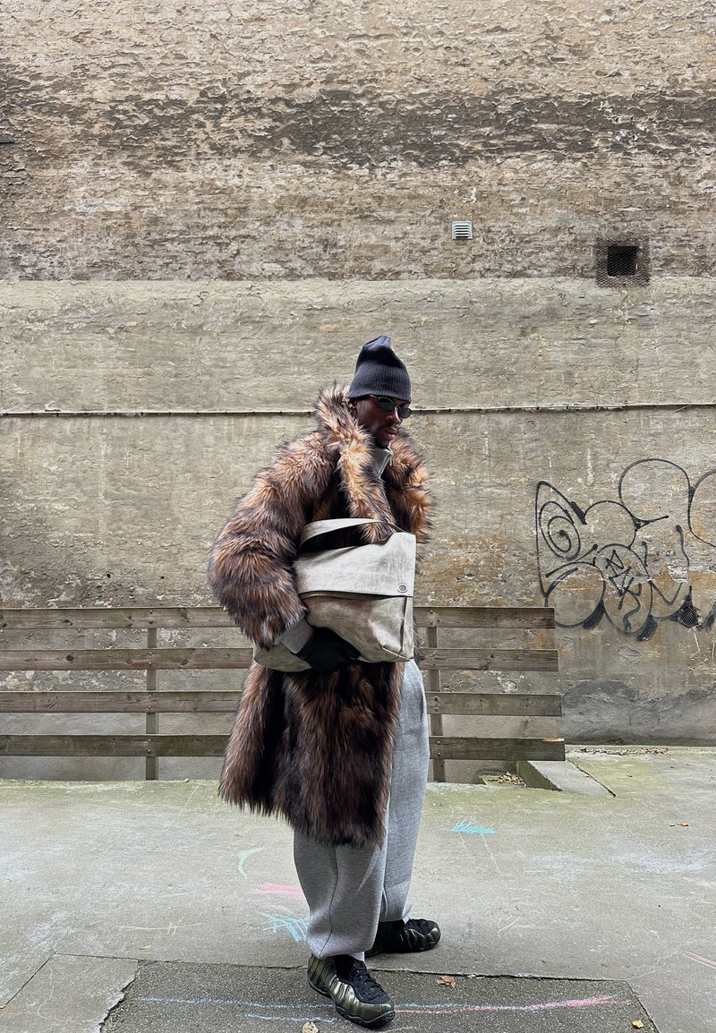 Manteau en fausse fourrure marron à manches longues, pantalon de survêtement gris et sac beige. Accessoires : bonnet noir et lunettes de soleil. Arrière-plan urbain.