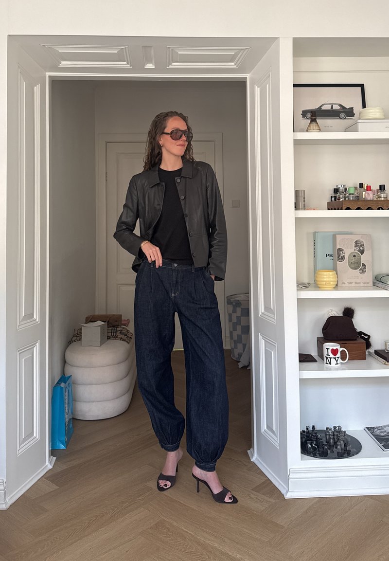 Femme portant une veste en cuir noire, un pantalon sombre ample, des sandales noires à talons ouverts et des lunettes de soleil, debout dans une embrasure de porte blanche à l’intérieur d’une pièce.