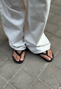 Chinelos pretos com uma sola texturizada e tiras em forma de Y, usados com calças brancas de corte solto, sobre uma superfície de azulejos cinzentos.