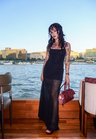 Vestido negro ajustado con paneles transparentes, combinado con medias de diseño. Los accesorios incluyen un bolso rojo con un colgante. Entorno de madera junto al agua.