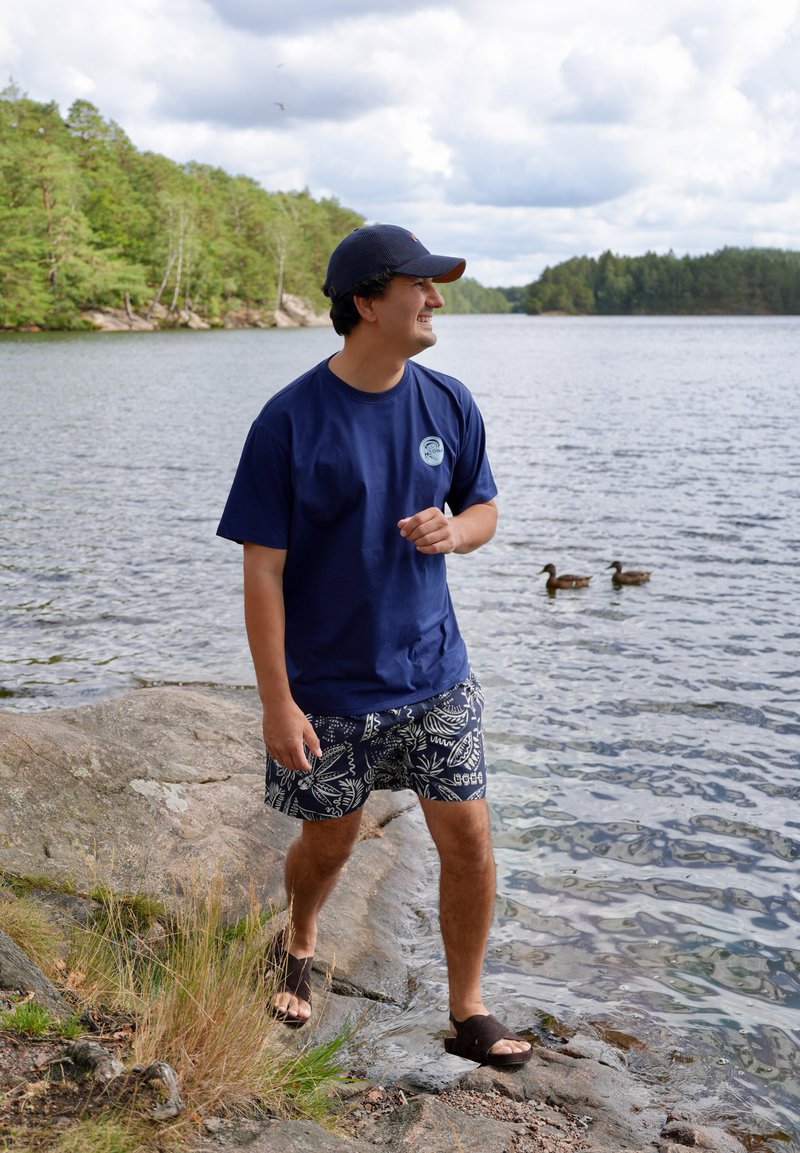 Young man wearing a navy shirt, patterned shorts, and cap walks barefoot on rocky lake shore with two ducks swimming nearby.