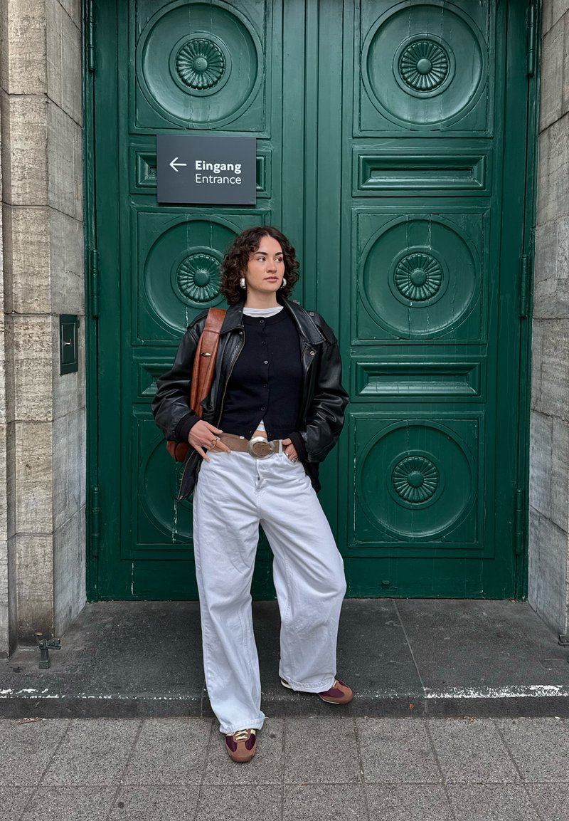 Jeune femme aux cheveux bouclés portant une veste noire et un pantalon blanc se tient près d'une grande porte verte avec un panneau "Entrée" en allemand et en anglais.