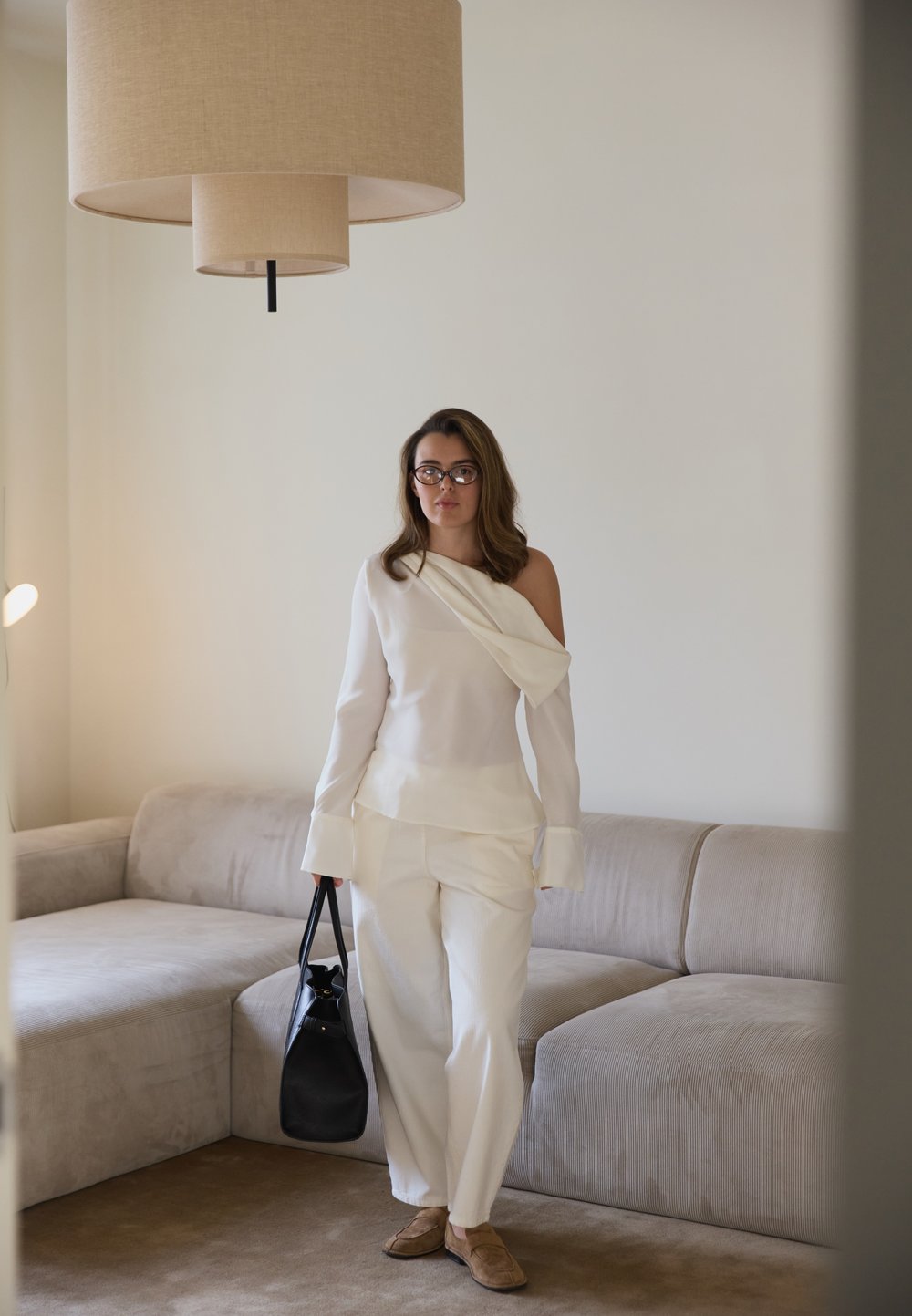 Bluză albă cu un singur umăr, din material drapat, asortată cu pantaloni albi largi. Poșetă neagră și pantofi bej confortabili. Fundal în tonuri neutre.