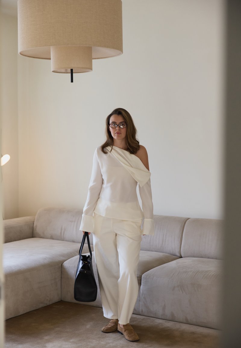 Weisses One-Shoulder-Blus mit drapiertem Material, kombiniert mit weissen, lockeren Hosen. Schwarze Handtasche und beige gemütliche Schuhe. Hintergrund in neutralen Tönen.