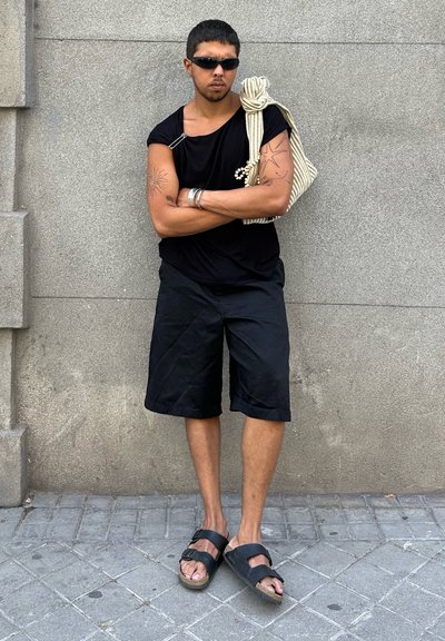 Camisa negra de manga corta con detalle de imperdible, pantalones cortos negros, sandalias negras y un bolso a rayas colgado del hombro. Fondo de pared gris.