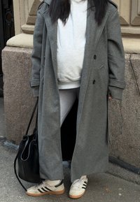 Personne portant un long manteau gris, un sweat à capuche gris clair, des leggings blancs, des baskets blanches avec des rayures noires, et tenant un sac à main noir.