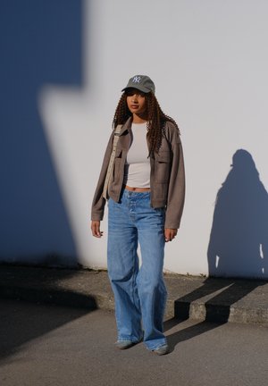 Una persona se encuentra de pie contra una pared blanca, vistiendo una gorra gris claro, una blusa crema, una chaqueta marrón de gran tamaño y unos vaqueros azules de pierna ancha.