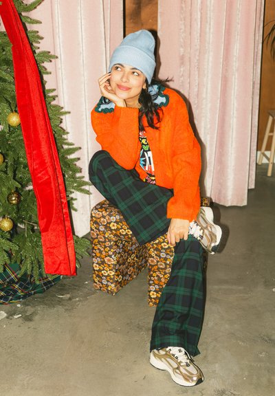Mujer con gorro azul, suéter naranja y pantalones de cuadros sentada en un cubo floral cerca de un árbol de Navidad con una cinta roja, apoyando la barbilla en la mano y sonriendo.