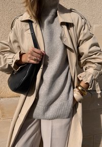 Beige trenčkot přes světle šedý pletený svetr s vysokým límcem. Černá kožená taška a šperky zahrnují zlatý náramek a prsteny.