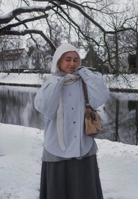 Femme en vêtements d'hiver avec une écharpe blanche et un collier de perles, se tenant près d'une rive enneigée avec des arbres nus et des maisons en arrière-plan.
