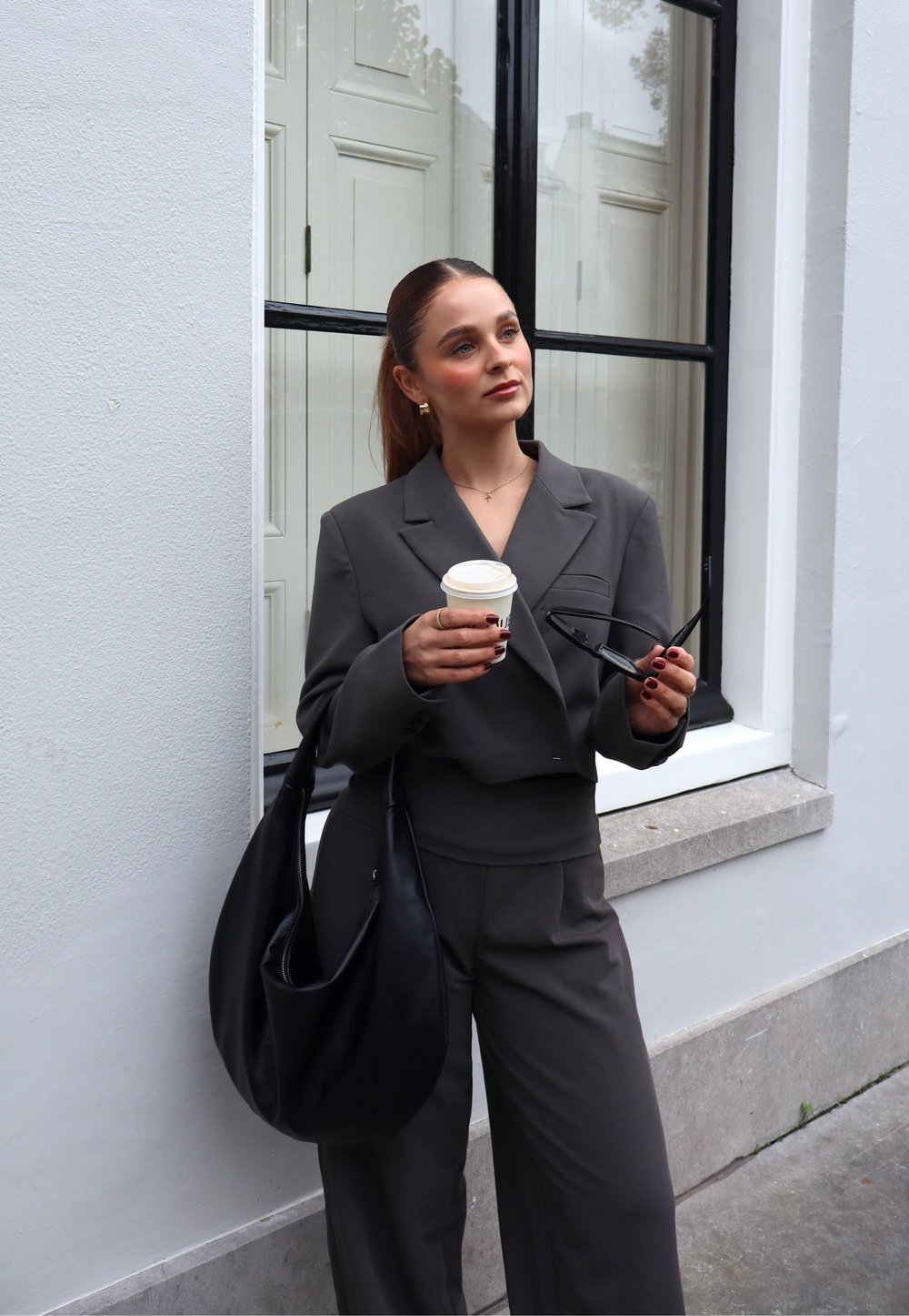 Γκρι κομμένο σακάκι με κοντή σχεδίαση, ταιριαστά παντελόνια σε φαρδιά γραμμή, μια μαύρη δερμάτινη τσάντα, κρατώντας ένα φλιτζάνι καφέ και γυαλιά.