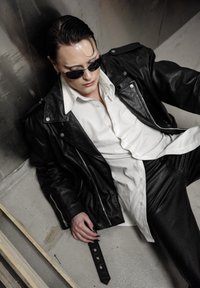 Veste en cuir noire sur une chemise blanche, pantalon en cuir noir, et lunettes de soleil. Accessoires comprennent une sangle noire avec des boutons-pression.