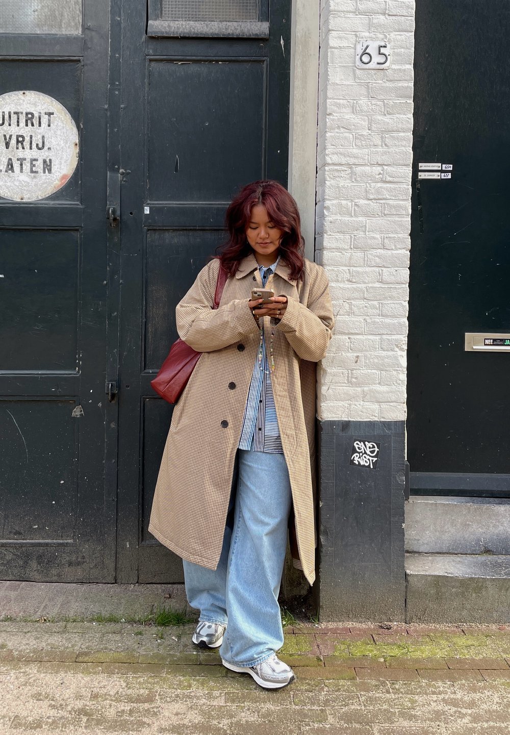 Beige hundtannjakke, løse lyseblå jeans, stripete skjorte og grå joggesko. Brun skulderveske. Svart dør og murvegg i bakgrunnen.