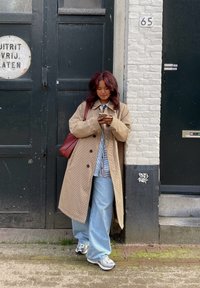 Beige hundtannjakke, løse lyseblå jeans, stripete skjorte og grå joggesko. Brun skulderveske. Svart dør og murvegg i bakgrunnen.