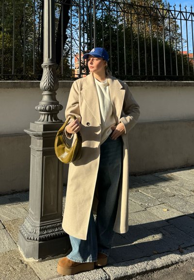 Abrigo de lana beige con cierre de doble botonadura, combinado con un suéter blanco y unos jeans azules de pierna acampanada. Lleva un bolso verde oliva y una gorra azul.