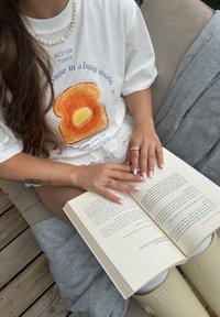 Hvit oversized t-skjorte med et toast-trykk, lagde blondeshorts, åpen bok med tekst, og kremfargede gummistøvler på en teksturert overflate.