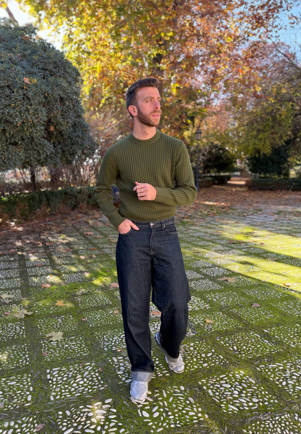 Homme portant un pull vert olive et un jean foncé marchant sur un chemin de galets moussus dans un parc avec des arbres d'automne.