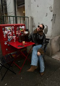 Femme en lunettes de soleil et veste en cuir assise à une table rouge en plein air, tenant une tasse blanche, avec du café et un téléphone sur la table près d'une boîte rouge couverte d'autocollants.
