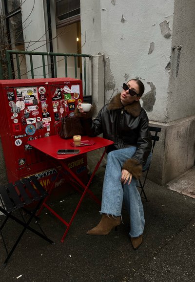 Mujer con gafas de sol y chaqueta de cuero sentada en una mesa roja al aire libre, sosteniendo una taza blanca, con café y teléfono sobre la mesa junto a una caja roja cubierta de pegatinas.