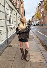 Bruine leren jas over een groene trui, zwarte mini-jurk en bruin kniehoge laarzen met crèmekleurige accenten. Stedelijke omgeving.