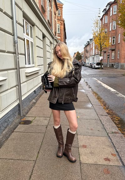 Chaqueta de cuero marrón sobre un suéter verde, falda corta negra y botas marrones hasta la rodilla con detalles en crema. Entorno urbano.