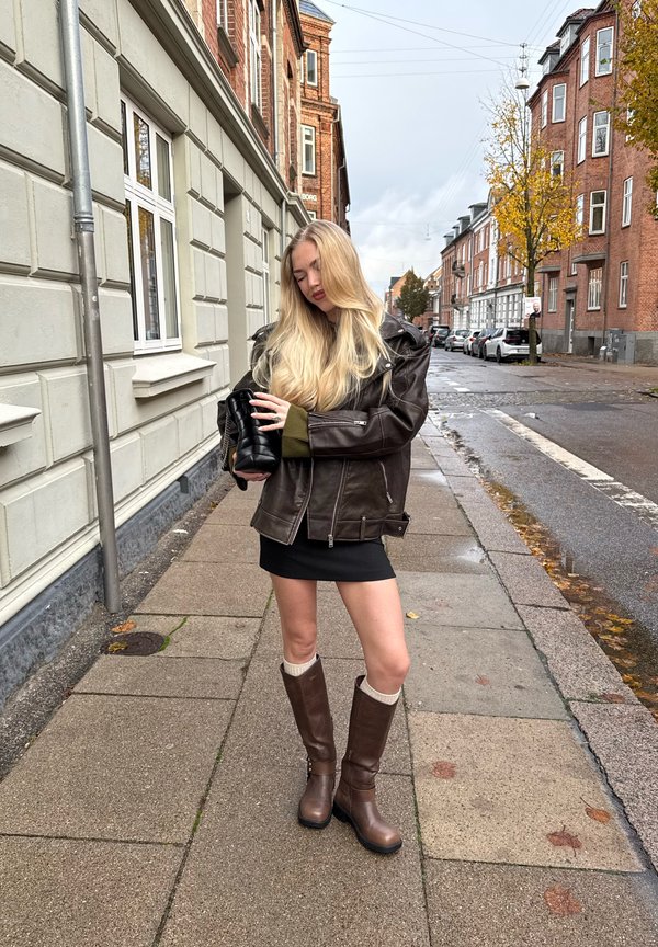 Hnědá kožená bunda přes zelený svetr, černá mini sukně a hnědé kozačky po kolena s krémovými akcenty. Městské prostředí.