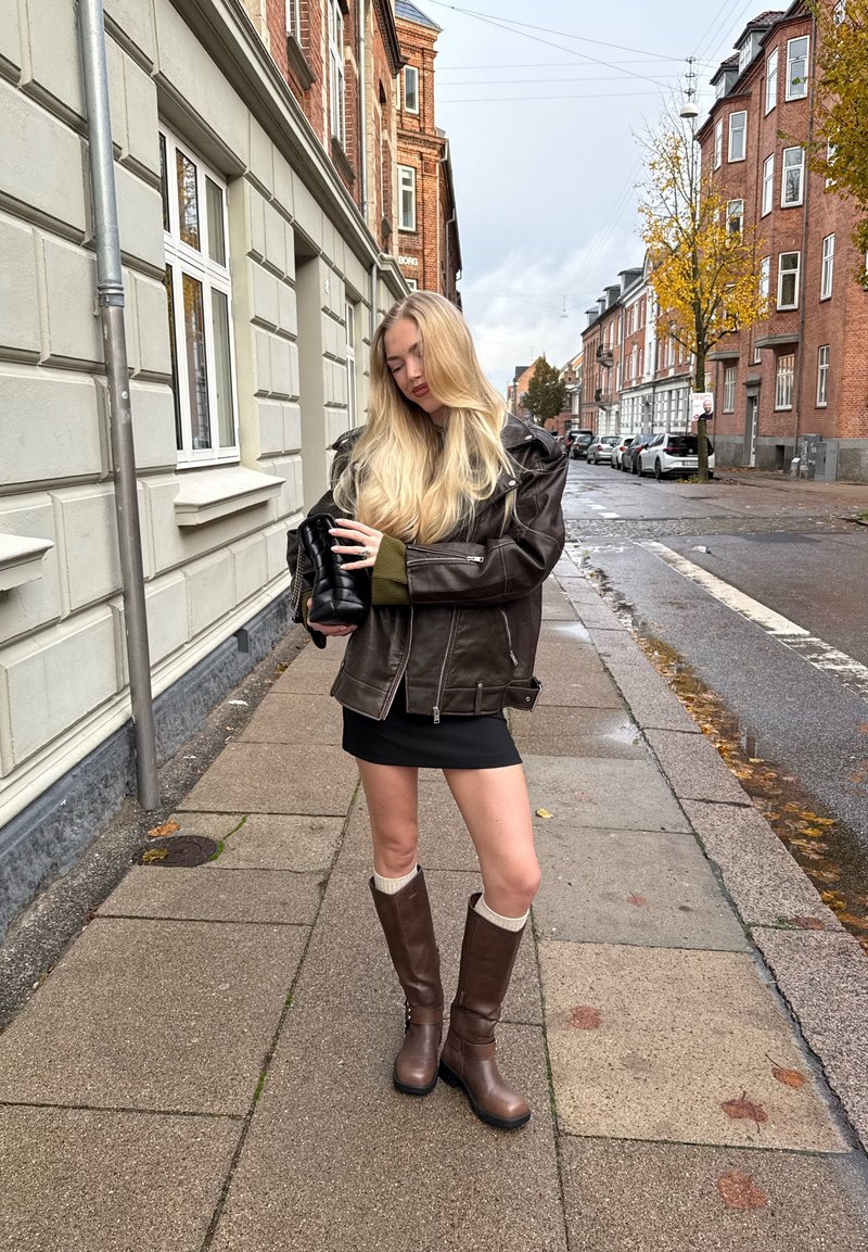 Smeđa kožna jakna preko zelene sweatshirto, crna mini suknja i smeđe čizme do koljena s krem detaljima. Urbana ulična postavka.