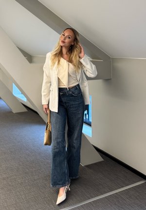 Mujer rubia con blazer blanco, top beige, vaqueros anchos oscuros y tacones blancos, de pie en un espacio interior moderno sosteniendo un bolso marrón.