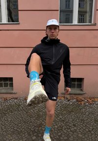 Zwarte sportjas met rits aan de voorkant, zwarte shorts, blauwe sokken en lichtgekleurde hardloopschoenen met gestructureerde zolen, tegen een roze muur.