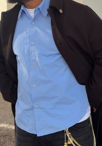 Camisa azul claro abotonada bajo un abrigo negro; jeans de mezclilla; cadena con cuentas y adornos sujeta en la cintura. Textura suave, diseño casual.