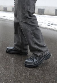 Άτομο που φοράει φορέματα με μαύρα παπούτσια με χοντρή σόλα και φαρδιά γκρι τζιν, στέκεται σε βρεγμένο πεζοδρόμιο κοντά σε λιωμένο χιόνι.