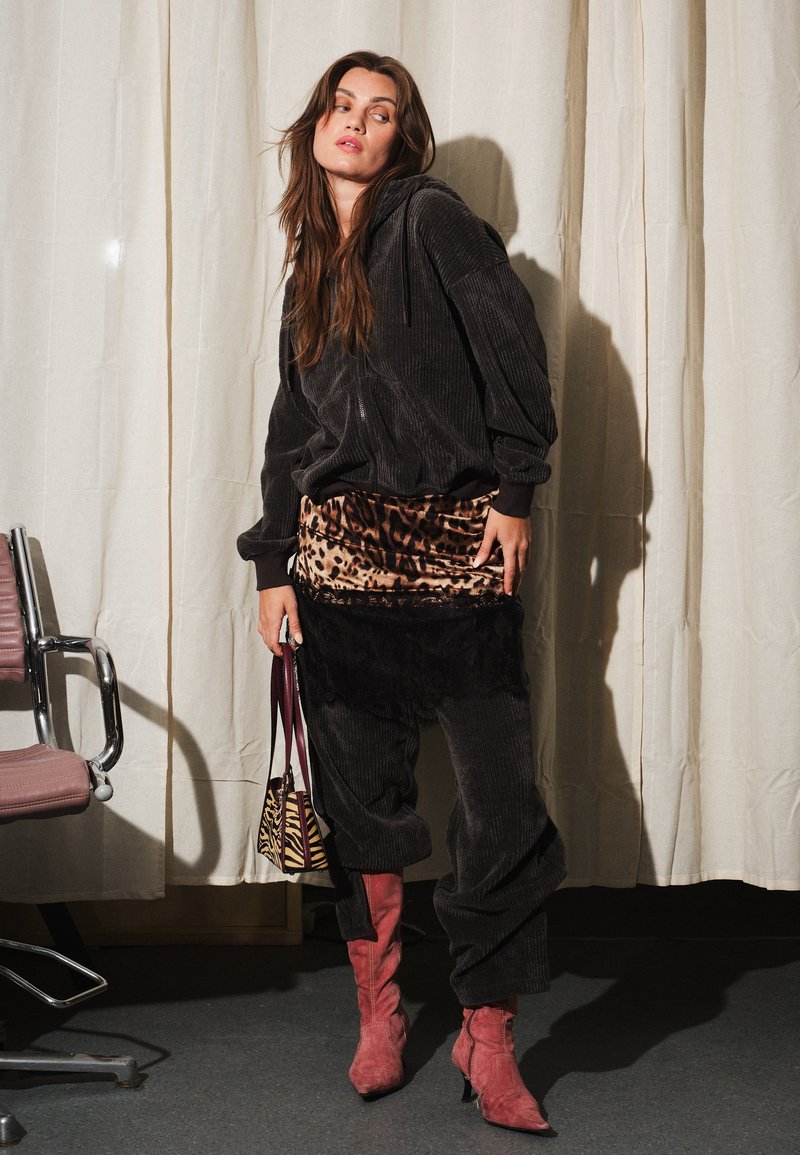 Femme debout, tête tournée, portant un sweat à capuche en velours côtelé noir et un pantalon, avec un par-dessus jupe imprimé léopard, des bottes rouges, et tenant un sac à main imprimé zèbre.