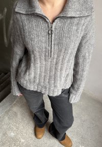 Grey knitted jumper with a half-zip collar, featuring a ribbed texture. Paired with black trousers and brown ankle boots.