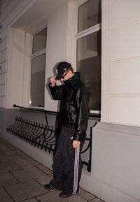 Veste en cuir noire à col montant, pantalon noir à motifs avec des rayures blanches, casquette de baseball et lunettes de soleil portés dans un cadre urbain.
