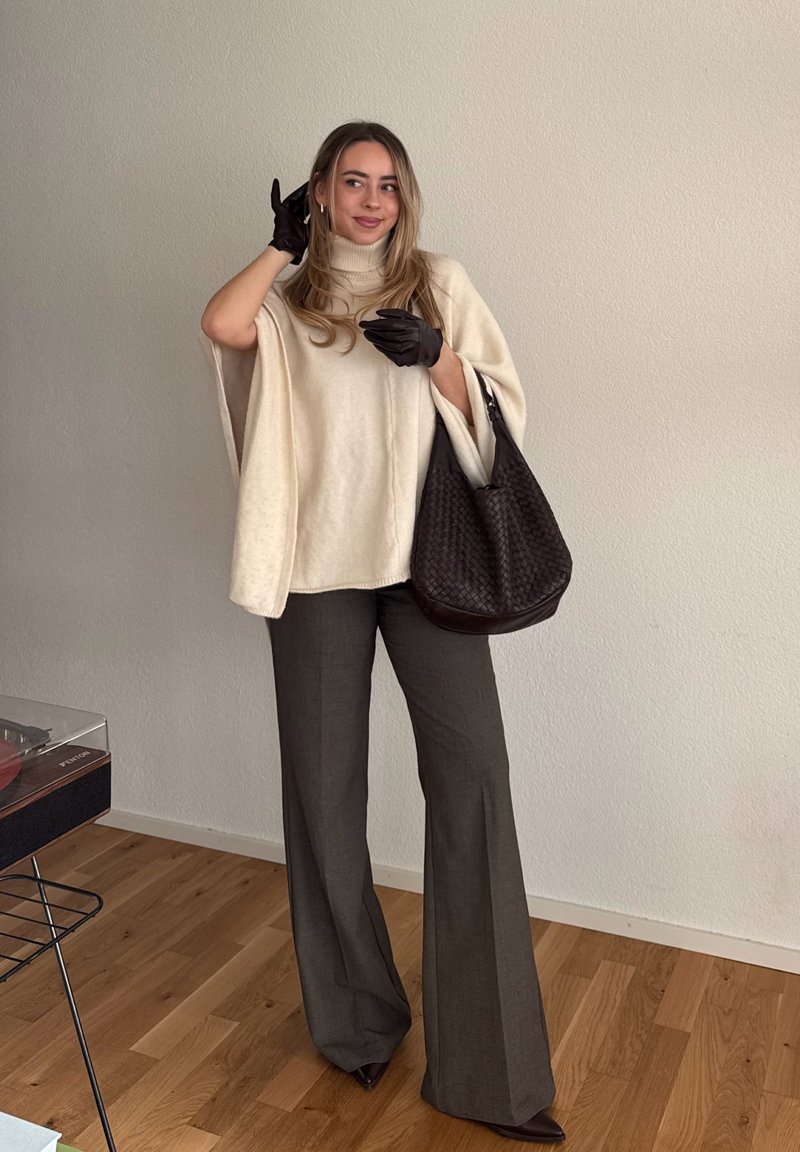 Kobieta stoi w pomieszczeniu, ubrana w kremowy sweter z peleryną, ciemne szerokie spodnie, czarne rękawiczki i trzyma dużą ciemną plecioną torbę.