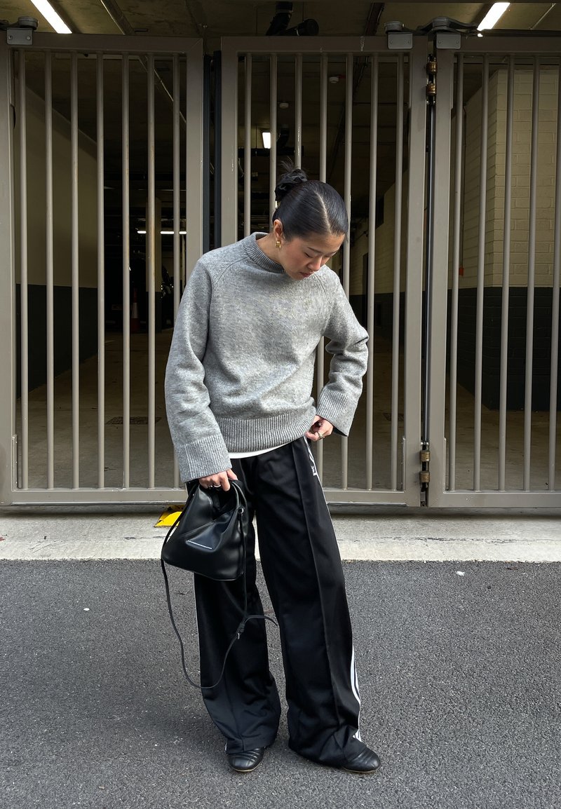Pull gris texturé à col montant ; pantalon noir à jambes larges avec bandes latérales ; tenant un petit sac noir ; debout dans un garage ouvert.