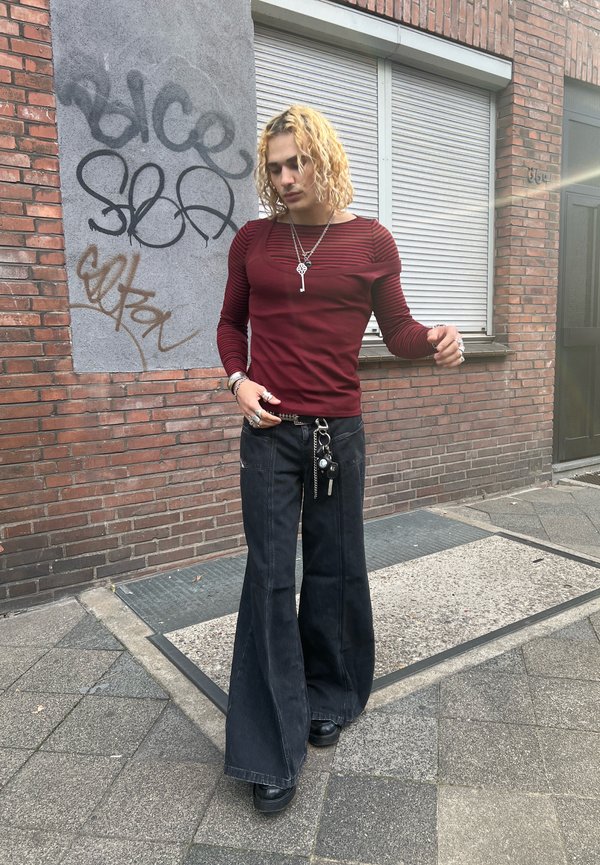 Maglietta rossa a maniche lunghe con righe orizzontali, abbinata a pantaloni in denim neri a gamba larga. Gli accessori includono catene e più anelli.