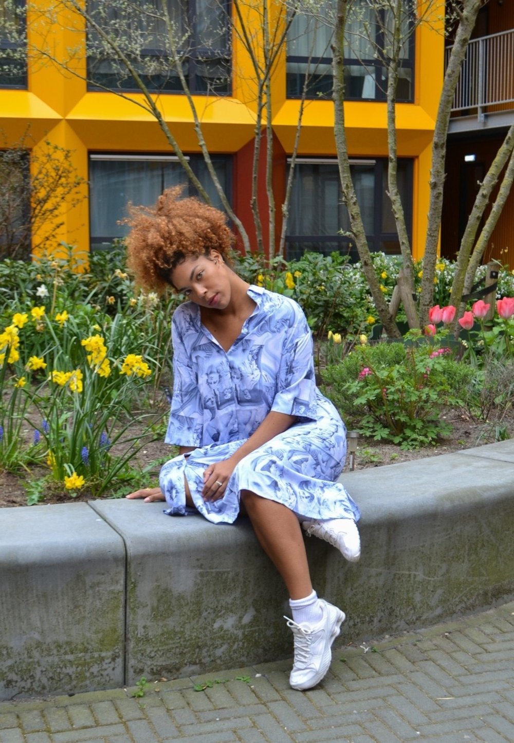 Robe bleu clair avec des motifs abstraits, manches courtes et col, associée à des chaussures de sport blanches. Entourée de fleurs et de verdure.