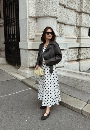 Mujer con chaqueta de cuero negra y vestido blanco con lunares, sosteniendo un bolso beige, de pie en la acera junto a un edificio de piedra texturizada.