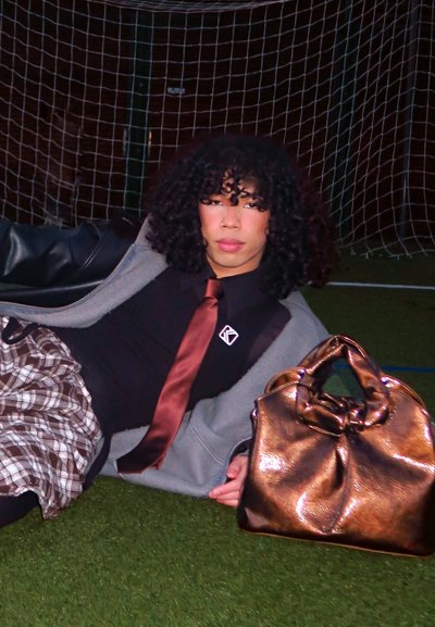 Bolso de mano brillante de bronce con asa retorcida, camiseta negra, falda de cuadros, chaqueta gris y corbata marrón, contra un fondo de césped verde con red de portería de fútbol.