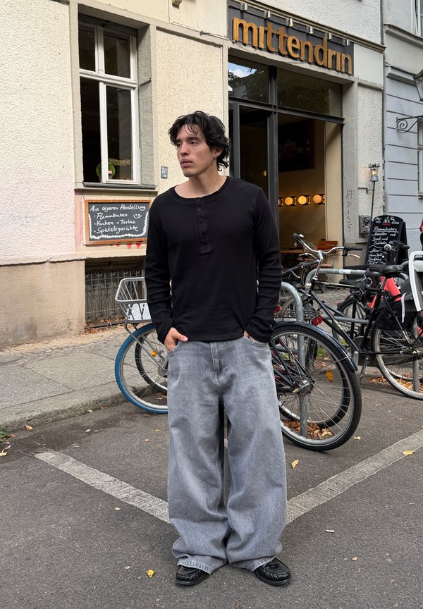 Mladý muž v čiernom tričku s dlhým rukávom a širokých sivých džínsoch stojí na ulici pri stojane na bicykle pred kaviarňou so znakom "mittendrin".
