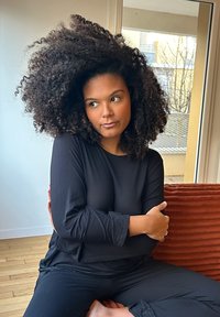 Maglietta nera a maniche lunghe e pantaloni neri, realizzati in tessuto testurizzato. Il soggetto ha capelli voluminosi e ricci, seduto con le braccia incrociate su un cuscino di tessuto arancione.