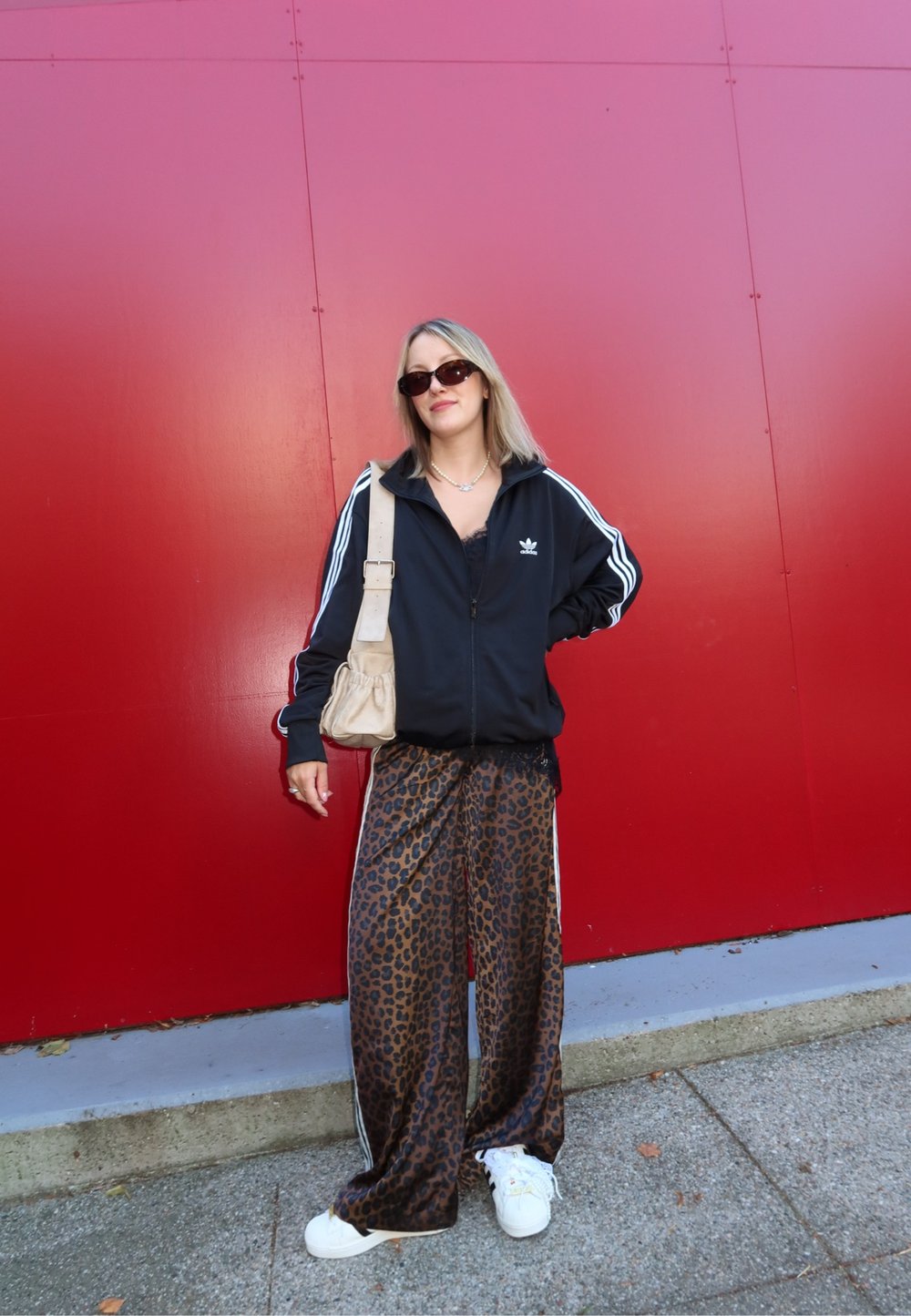 Jachetă neagră cu fermoar, cu dungi albe, pantaloni largi cu imprimeu de leopard și adidași albi. Ține în mână o geantă bej, stând de-a lungul unui perete roșu.