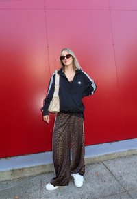Jachetă neagră cu fermoar, cu dungi albe, pantaloni largi cu imprimeu de leopard și adidași albi. Ține în mână o geantă bej, stând de-a lungul unui perete roșu.